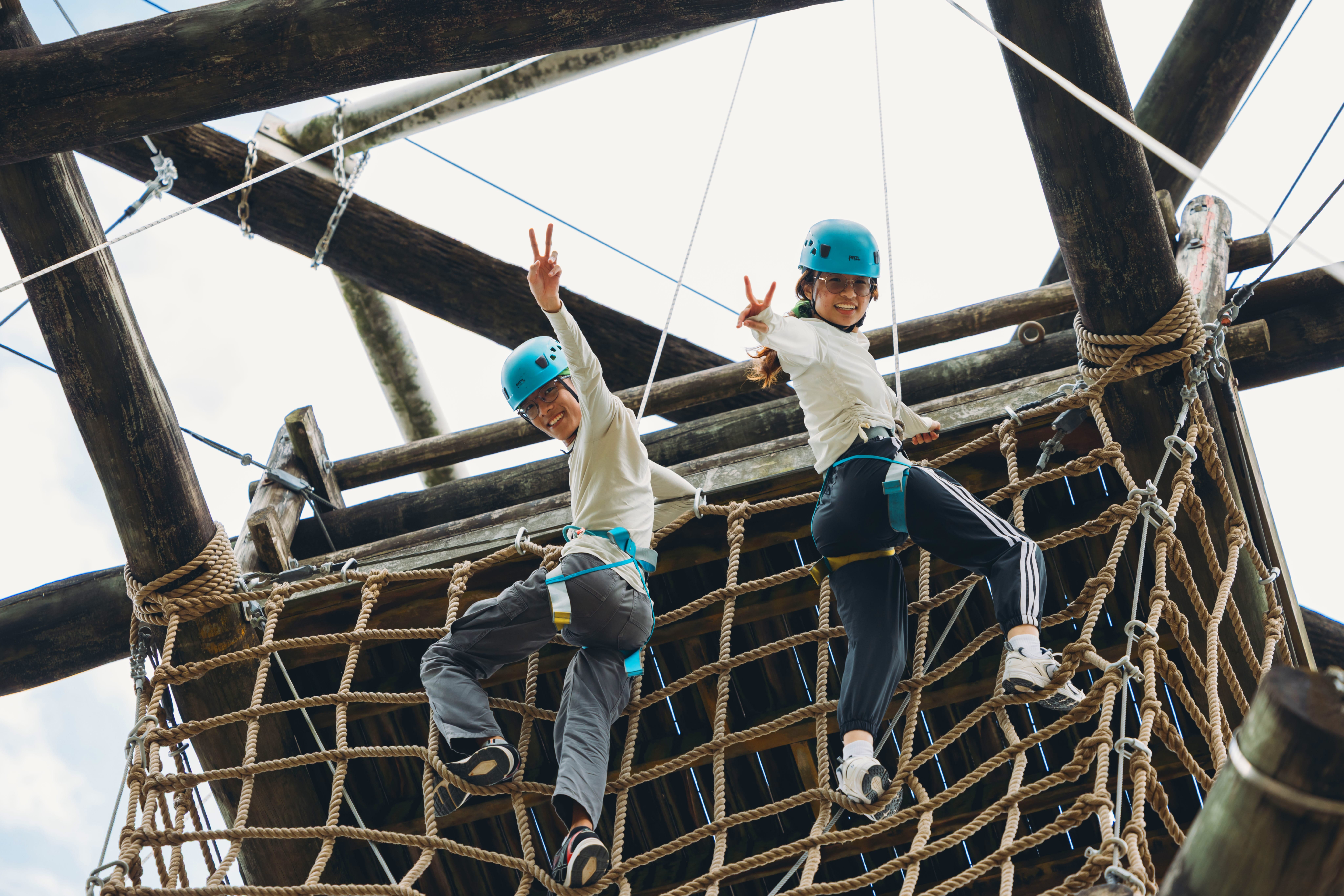 Two people in helmets and safety harnesses climb a rope net on a wooden structure.